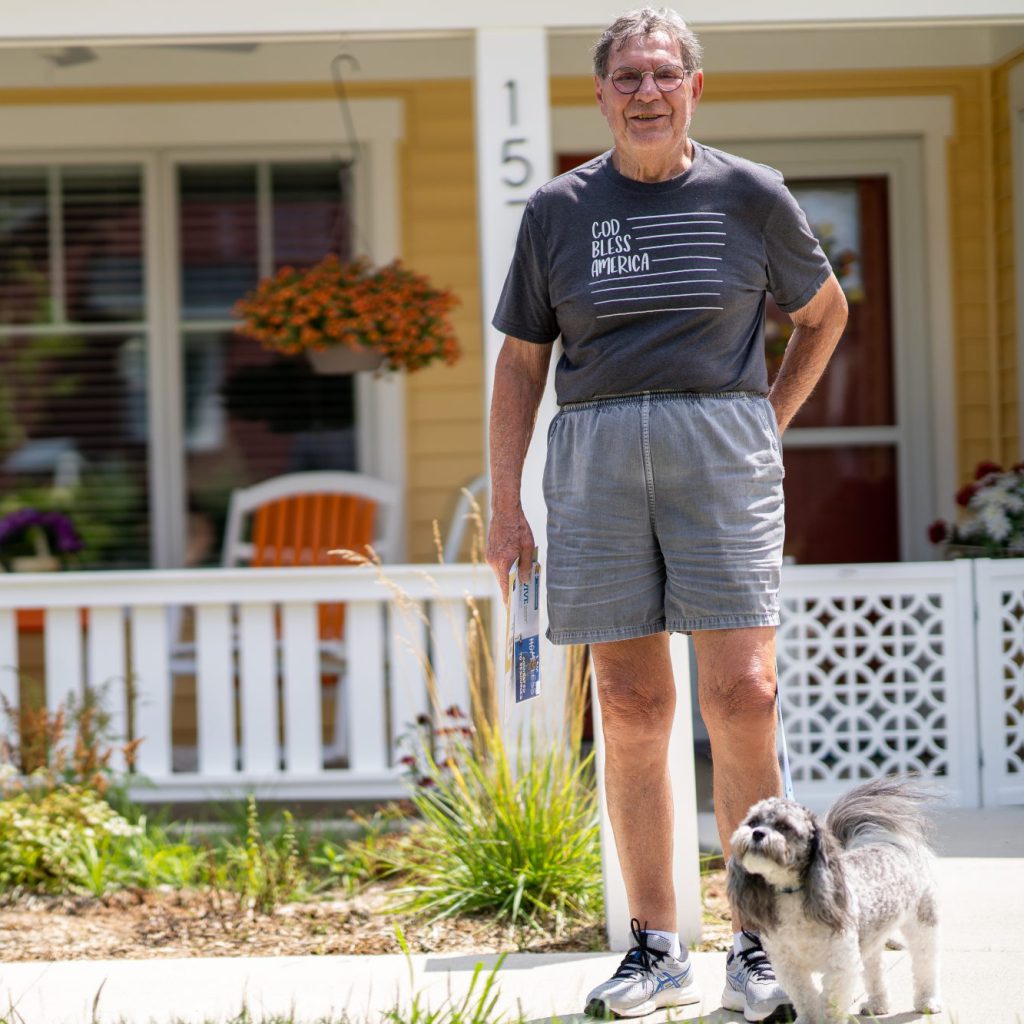 An older gentleman stands in front of the front porch of a charming yellow cottage. He looks warmly at the camera and is holding mail, and he wears a gray shirt with the text "God bless America." In front of him, a small gray-and-white dog on a leash.