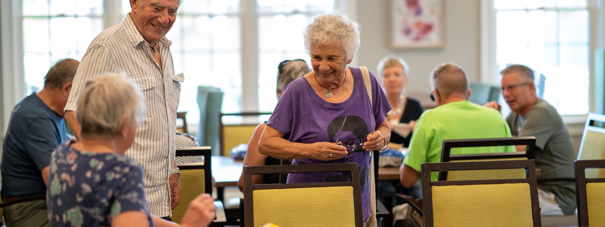 In the background, people are seated around a table. In the foreground, an older woman and man are stopped to speak with another woman with food at a table. They all are smiling.