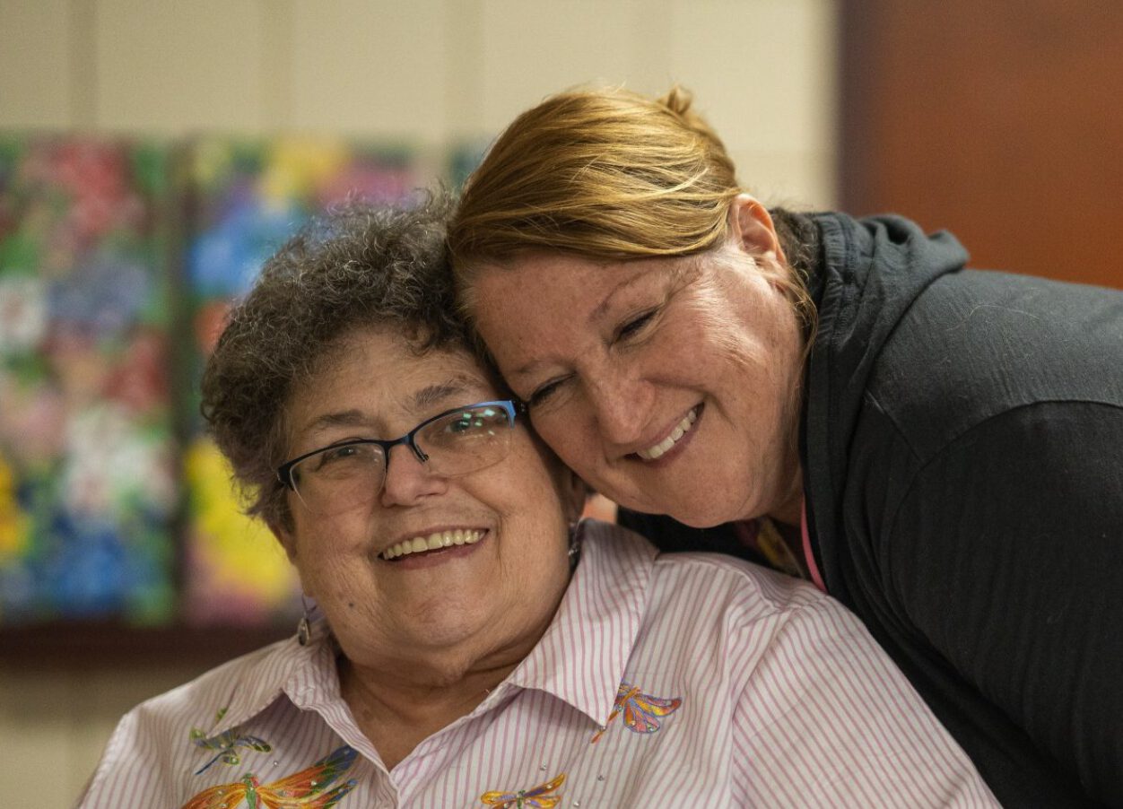 Two women, one sitting and one standing, embrace closely while smiling at the camera. The woman sitting is slightly older and wears a pink striped dragonfly shirt and glasses. The woman standing is a bit younger and has brown hair and freckles.